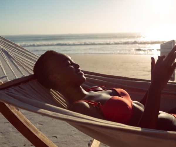 Side view of happy African-american woman using digital tablet while relaxing in a hammock on the beach